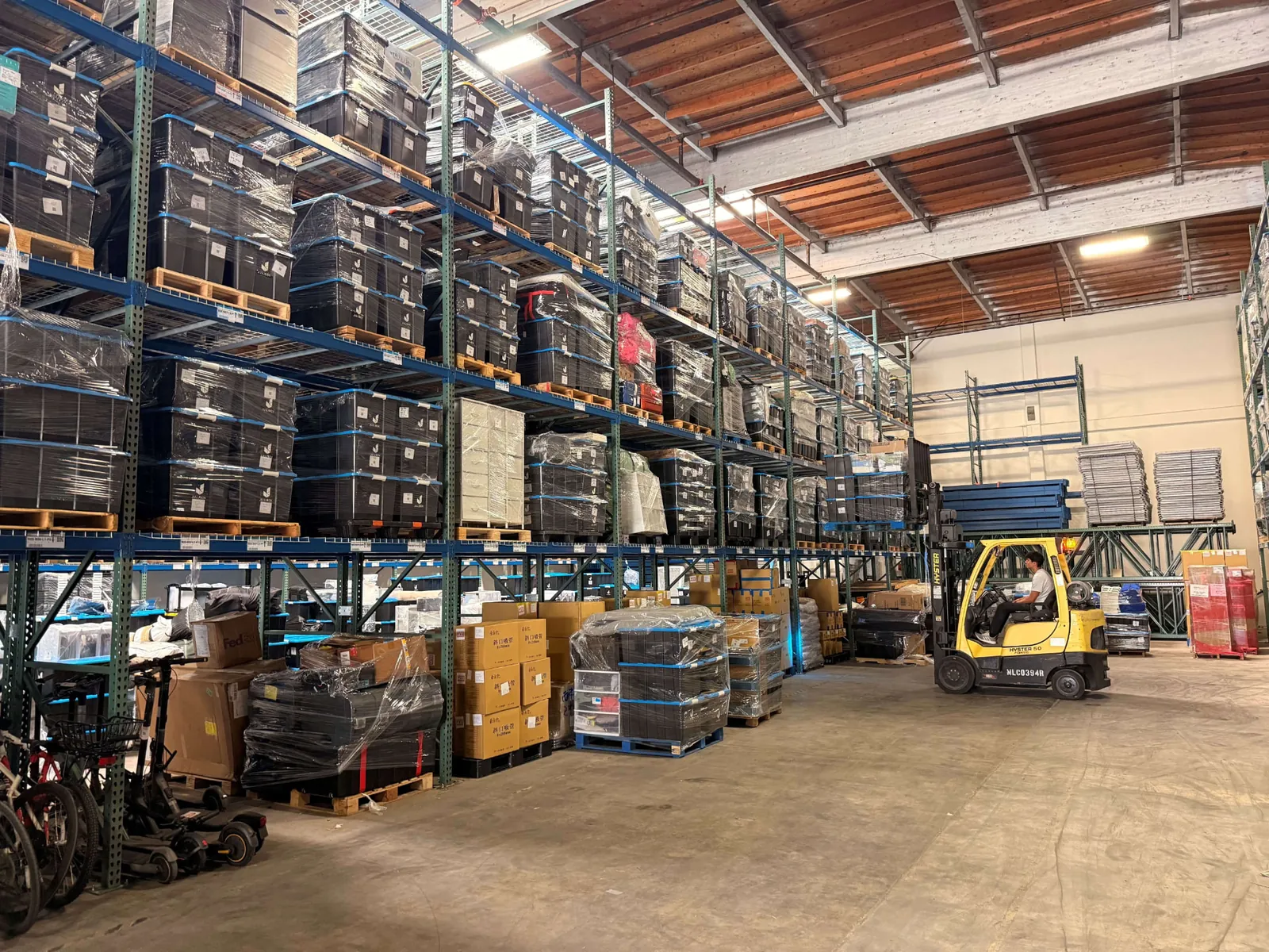 Warehouse racking loaded with shrink-wrapped bins, totes, and boxes, with a forklift driving the aisle